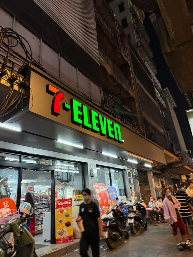 חזית מוארת של חנות 7-Eleven ברחוב בבנגקוק בלילה עם קטנועים חונים בחוץ